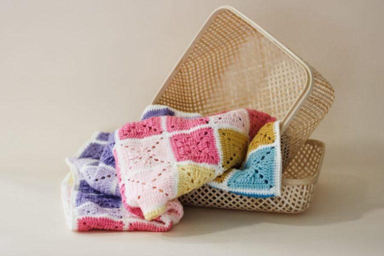 Colorful handmade crochet blanket made from granny squares in pink, purple, yellow, and blue tones, draped over a woven basket on a neutral background