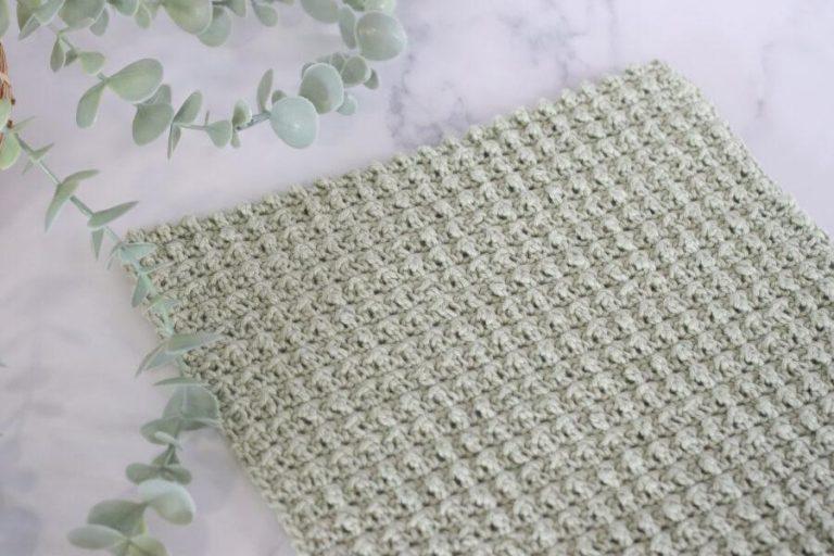 Close-up of a crocheted swatch using the cobble stitch in light sage green yarn, displayed on a marble surface with eucalyptus leaves in the background.