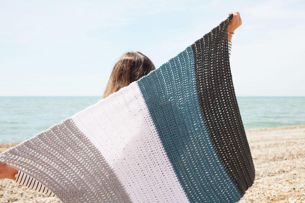 female holding up a striped crochet blanket in colours; grey, white and blue on a beach with the sea in the background. 
