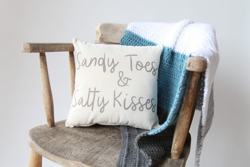 Crochet blanket on a wooden chair with a cushion that says 'Sandy toes & Salty Kisses' on it in the handwritten style.