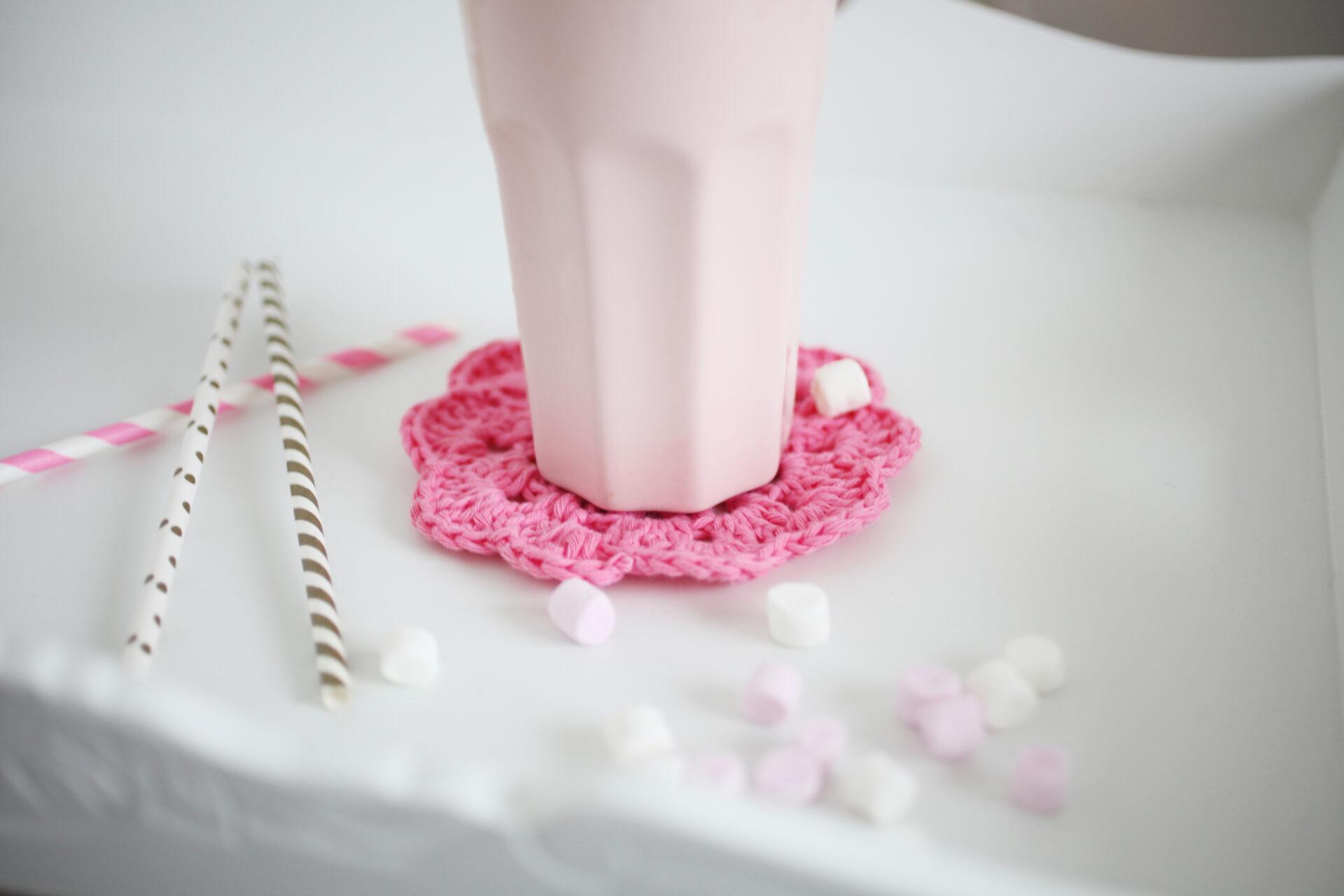 pink crochet coaster in a white tray surrounded by pink nd gold paper straws and mini marshmallows and a tall pale pink mug sat on the coaster