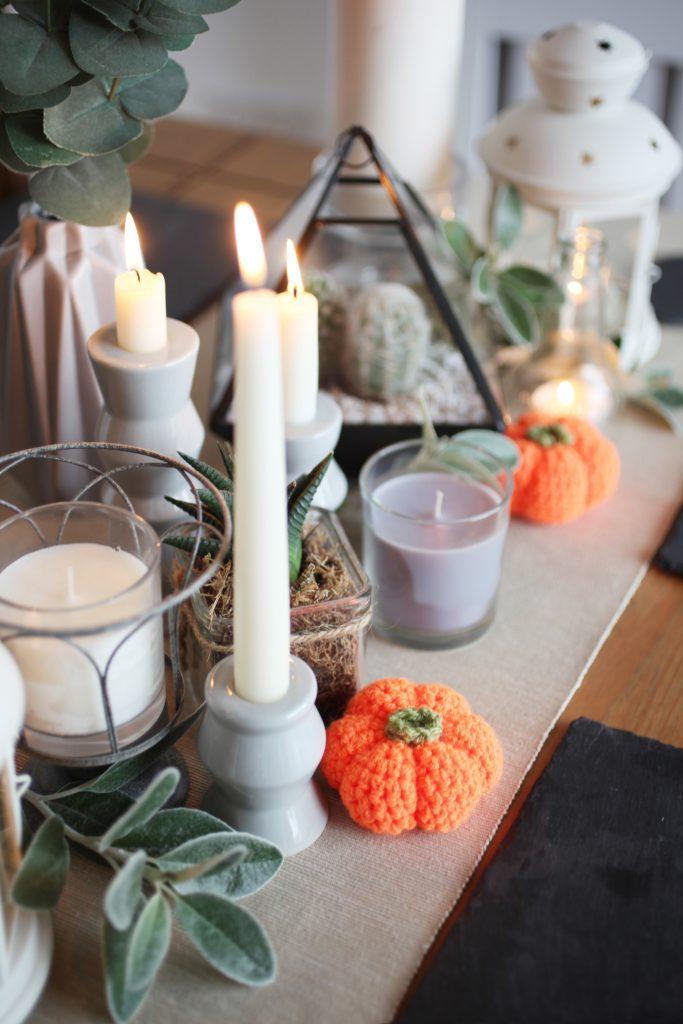 A table decoration with lots of mixed size candles, plants, foliage and two crochet pumpkins.
