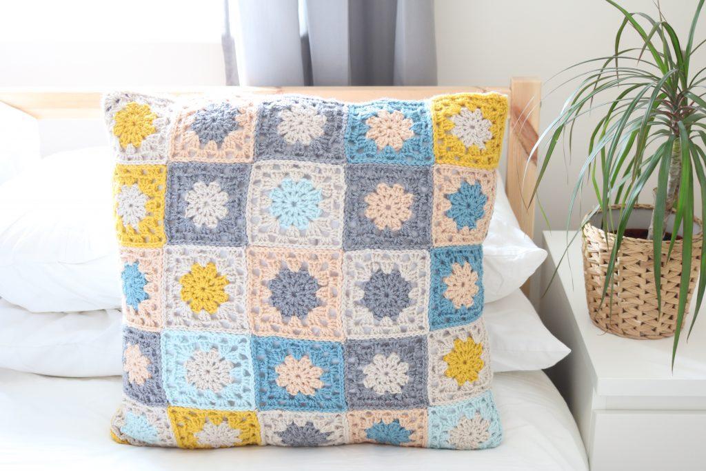 a granny square cushion made of mustard yellow, grey, pale blue and peach yarn sat on a white bed with a wooden frame. Next to the bed is a white bedside table with a plant sat in a wicker pot. 