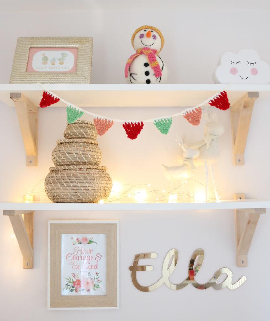 mini crochet bunting hanging from a white shefl in a little girls bedroom, the helfs are decorated with christmas ornaments and photo frames. The bunting is made in red, green and peach yarn attached together with cream yarn. 