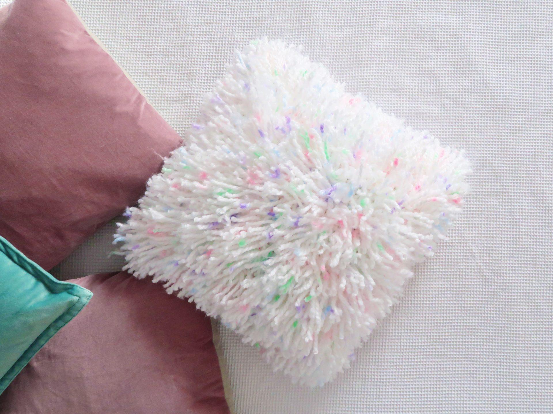 a flat lay image of a fluffy crochet cushion lay on cream carpet made of white yarn with specks of multi colours