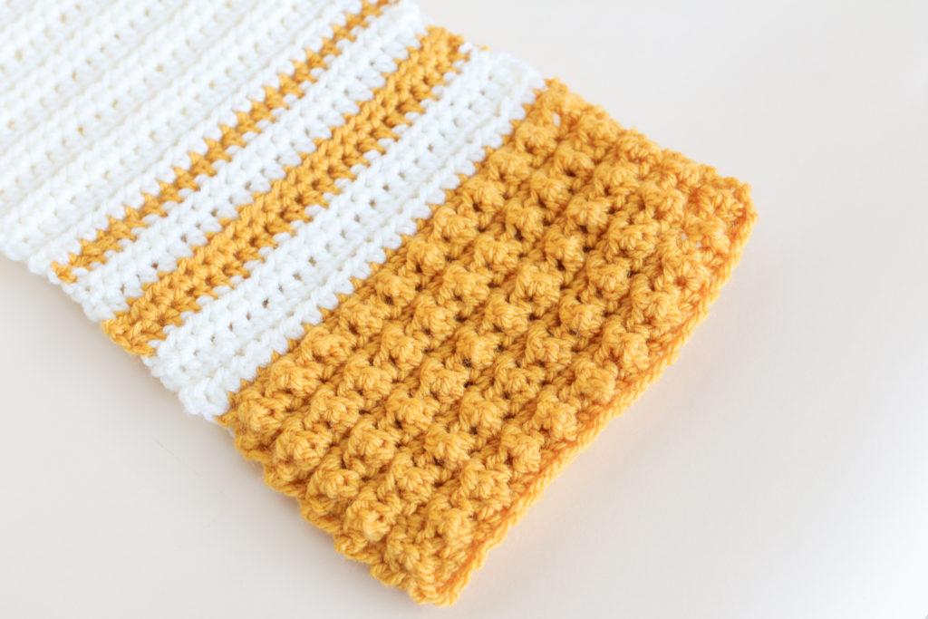 A yellow and white scarf made from crochet cobble stitch sits on a white background. 