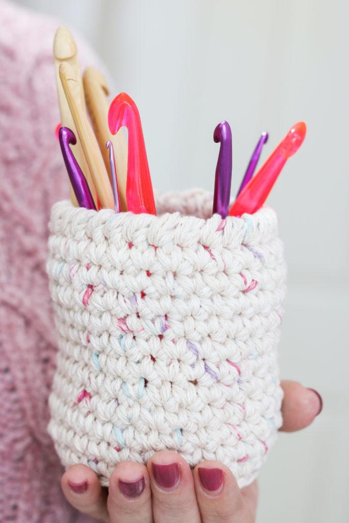 a woman's hand held out with a crochet storage pot in the palm of her hand. In the storage pot are a mixture of bamboo and plastic crochet hooks in different sizes and colours.
