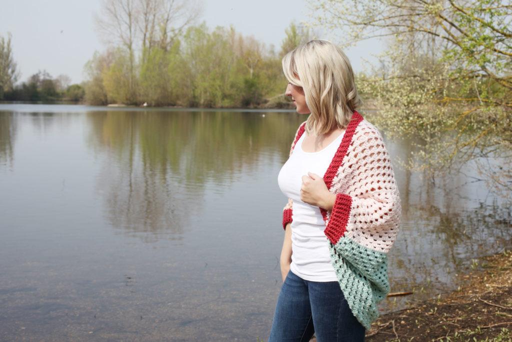 Sarah-Jayne stands in front of a lake, she is wearing a crochet cardigan in pastel colours with jeans and a white vest. She is looking to the side and smiling. 