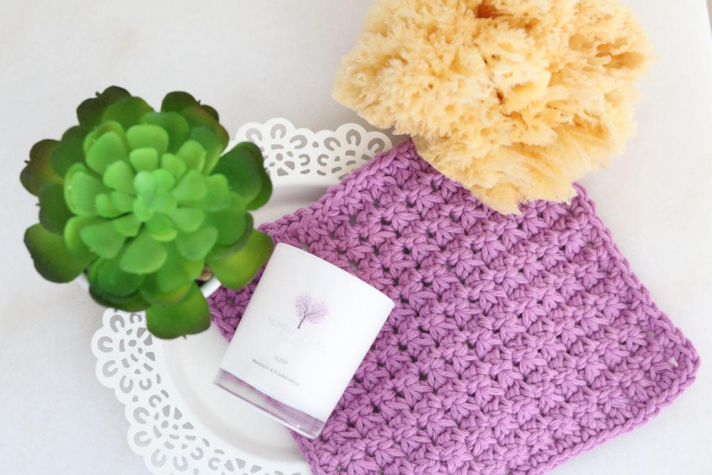 A crochet wash cloth with a candle and plant laid in a relaxing self care setting in a flatlay style.