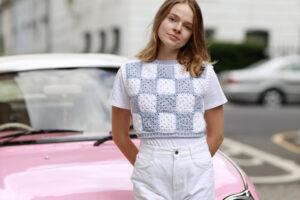 a female stood in front of a pink car in the street wearing white jeans and a crochet granny square t.shirt in white and blue granny squares.
