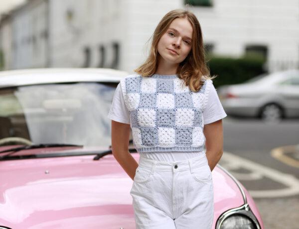 a female stood in front of a pink car in the street wearing white jeans and a crochet granny square t.shirt in white and blue granny squares.