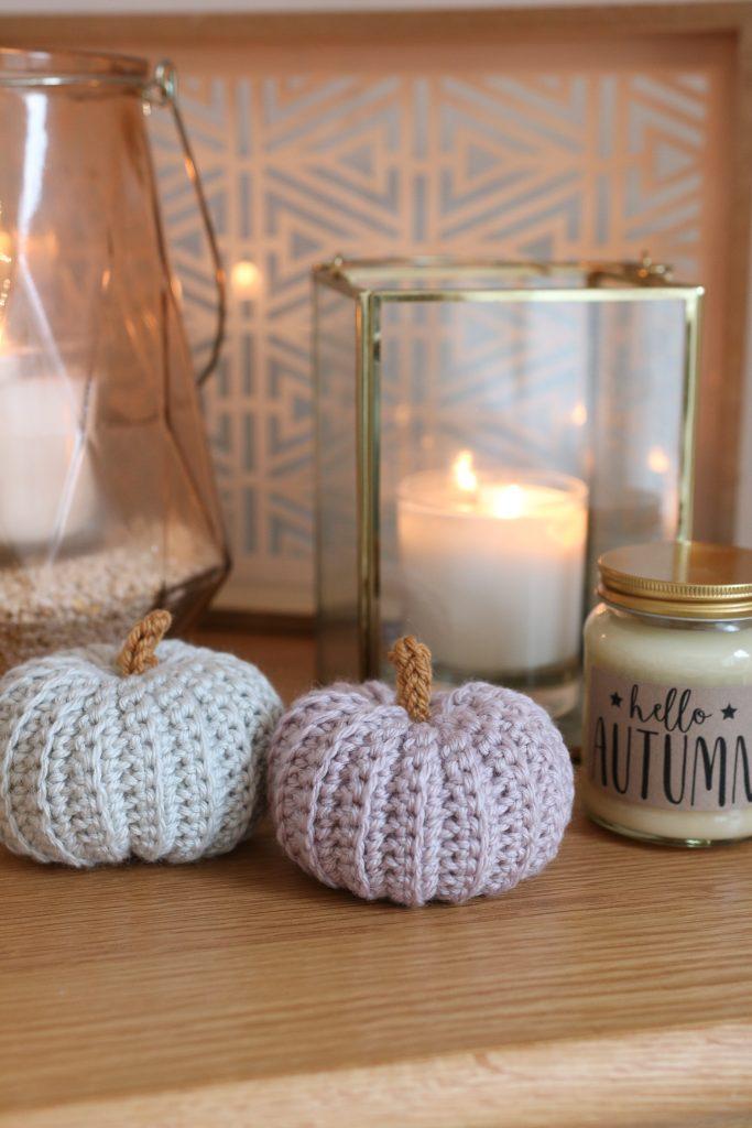 A pair of pastel-coloured crochet pumpkins sit on a fireplace in front of lit candles. 