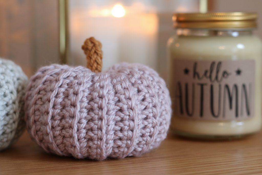 a pastel pink crochet pumpkin sits on a wooden surface beside a Autumn candle. 