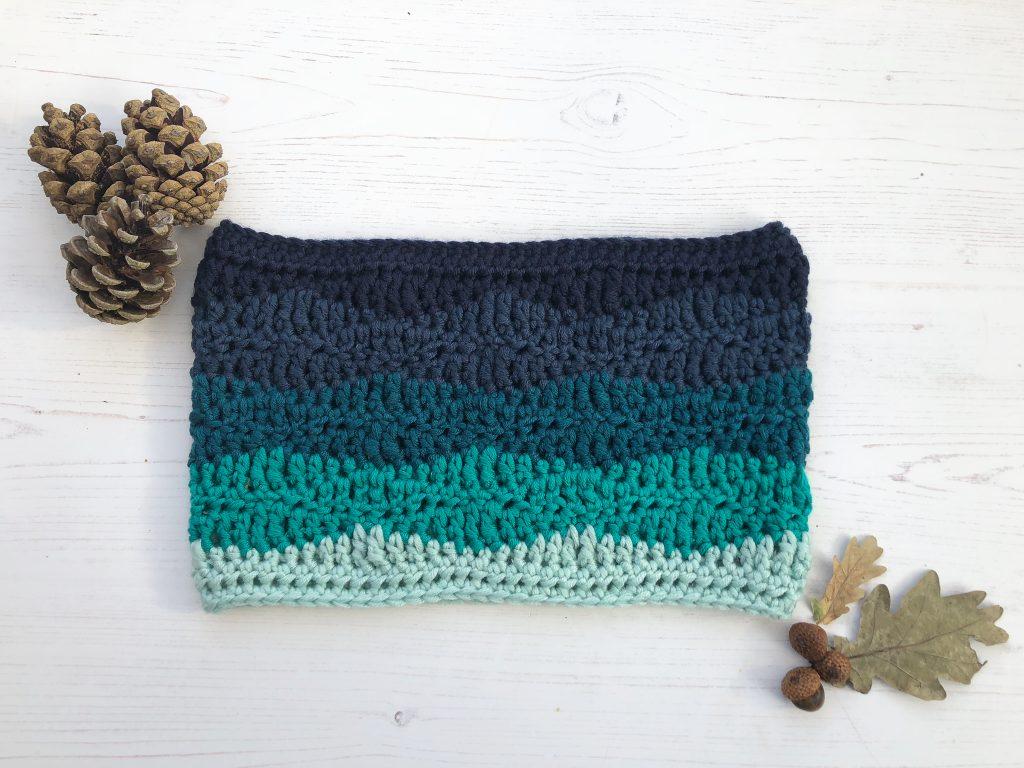 a flat lay image of a striped crochet cowl made in 5 different shades of blue going from light blue to navy blue, each section creates a wave design. The crochet cowl is lay on a white washed wooden backdrop with pine cones and leafs in the top and bottom corner of the flat lay. 