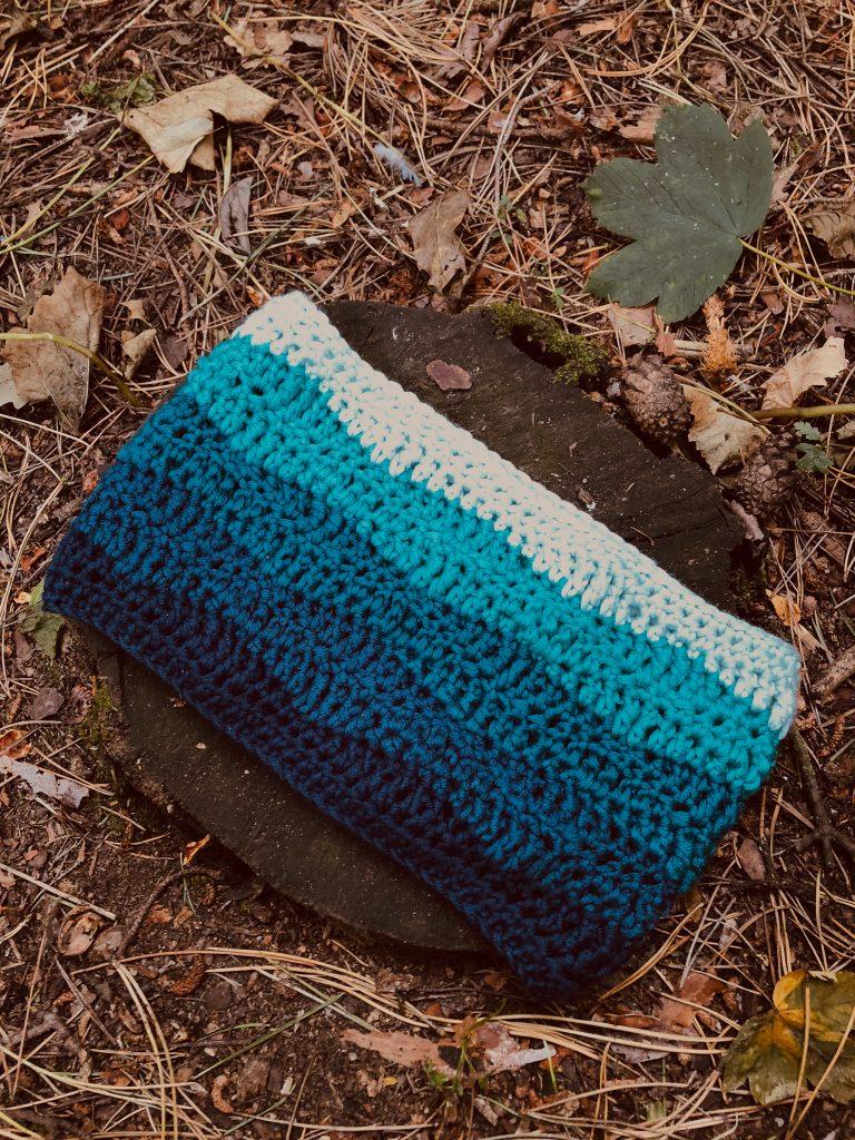 A cowl crocheted in 5 different types of blue in an ombre stripe look. The cowl is on a wooden plank in an outdoor setting surrounded by leaves and foliage like it's been left in the woods.