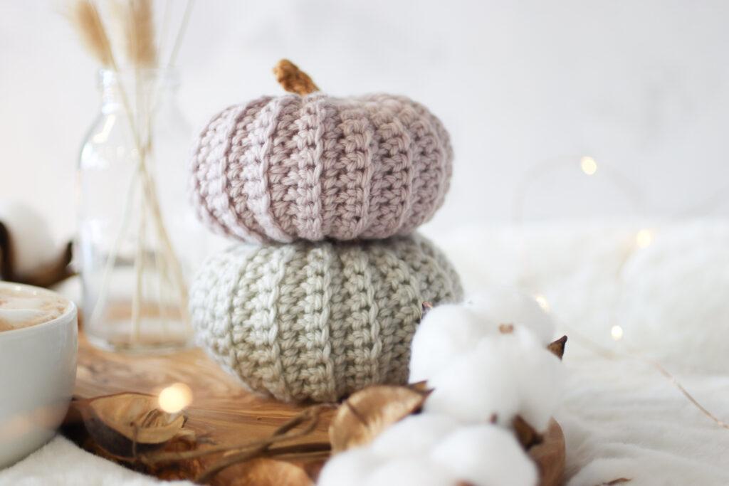 two crochet pumpkins stacked one grey and the other in mushroom. The crochet pumpkins are surrounded by small fairy lights with a cup of coffee and bunny tail dried flowers in a small glass vase. the background is white and the set up is placed on a white faux fur rug