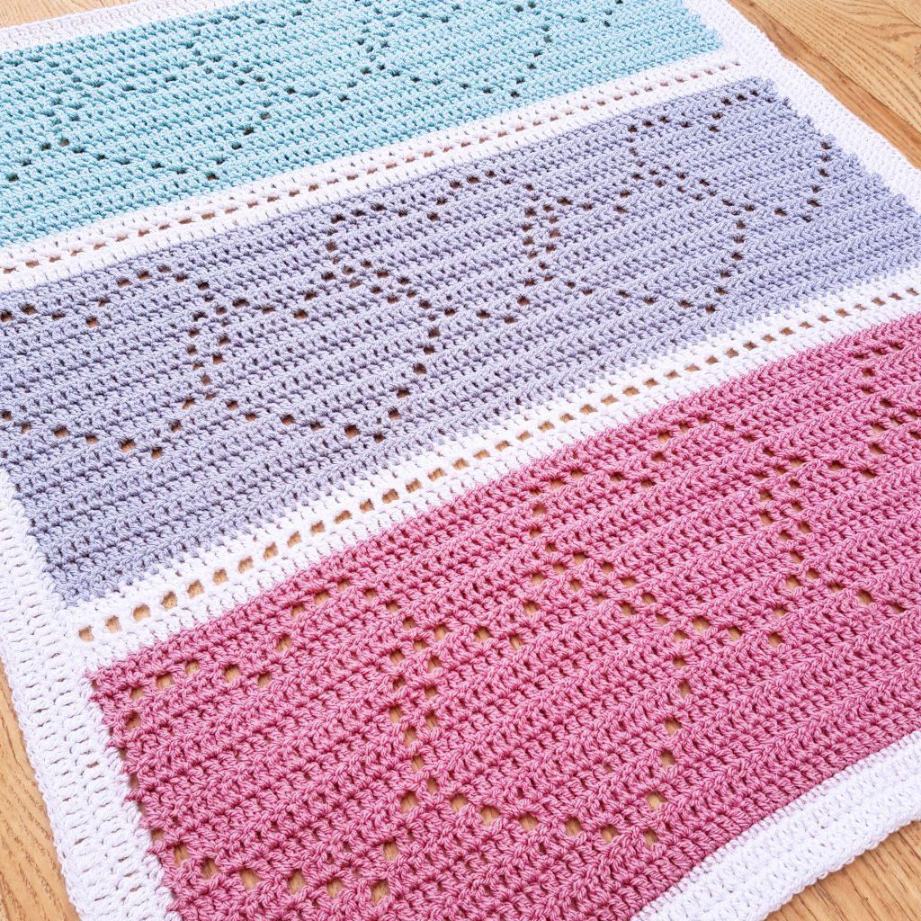 A filet crochet blanket in pastel shades of pink, blue and purple sits on a wooden background. 