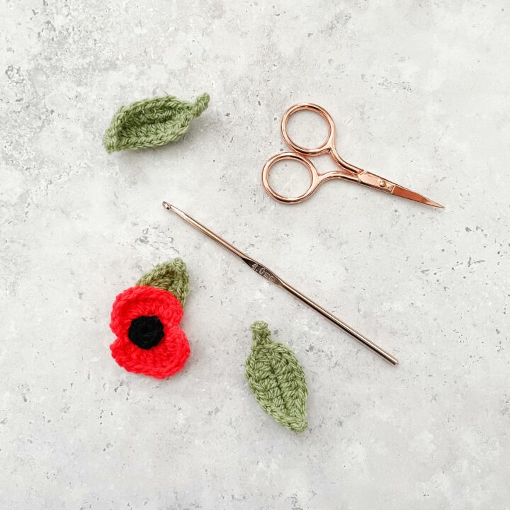 a flat lay image of a crochet poppy lay alongside two crochet leaves a rose gold hood and scissors lay on a concrete effect back drop.