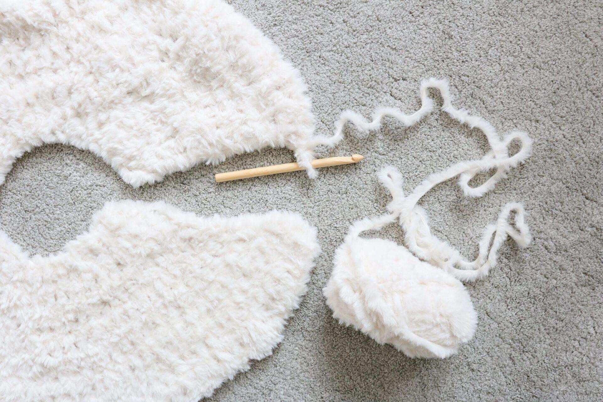 a flat lay image of a crochet tree skirt made from cream fluffy yarn lay on a beige carpet with the ball of yarn still attached and a wooden crochet hook. 