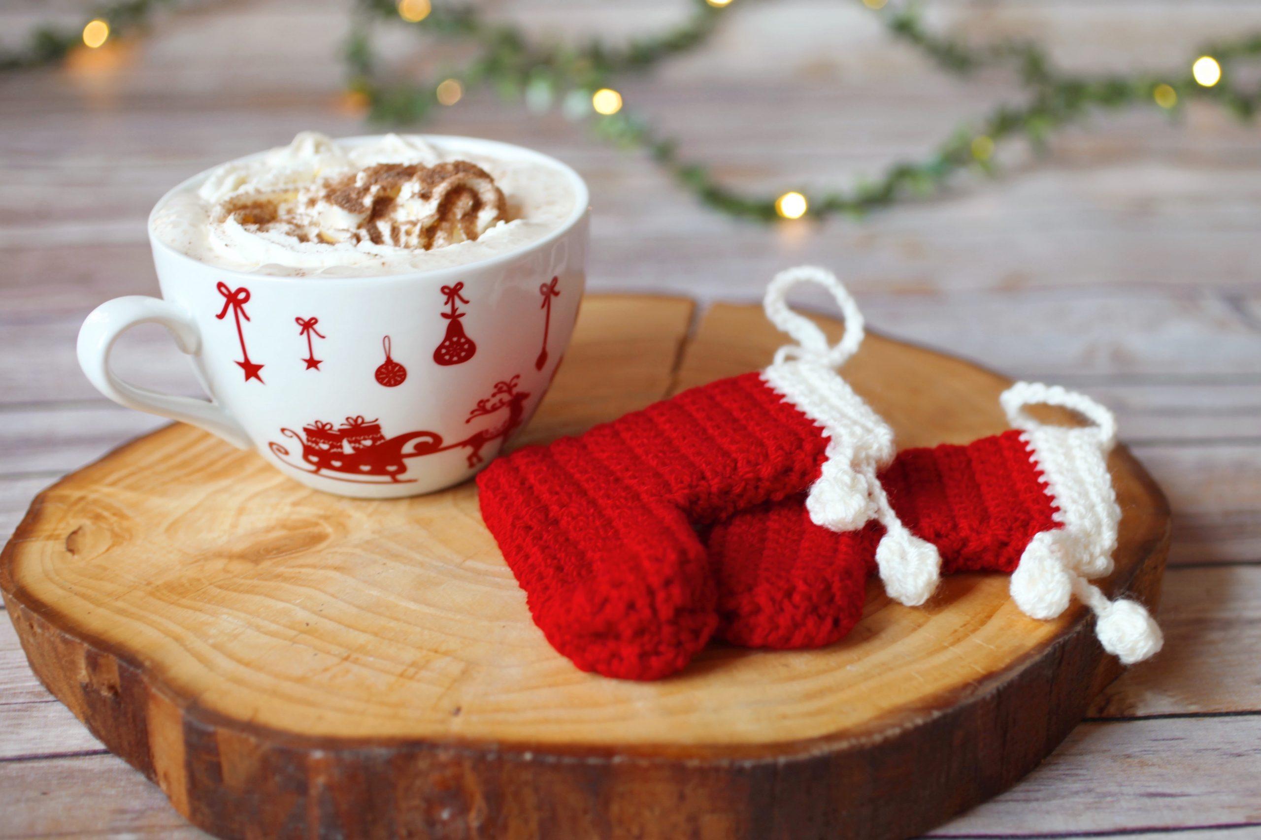 two Christmas Crochet Stockings made with red and cream yarn lay on a log slice alongside a Christmas mug filled with cream and chocolate sprinkles