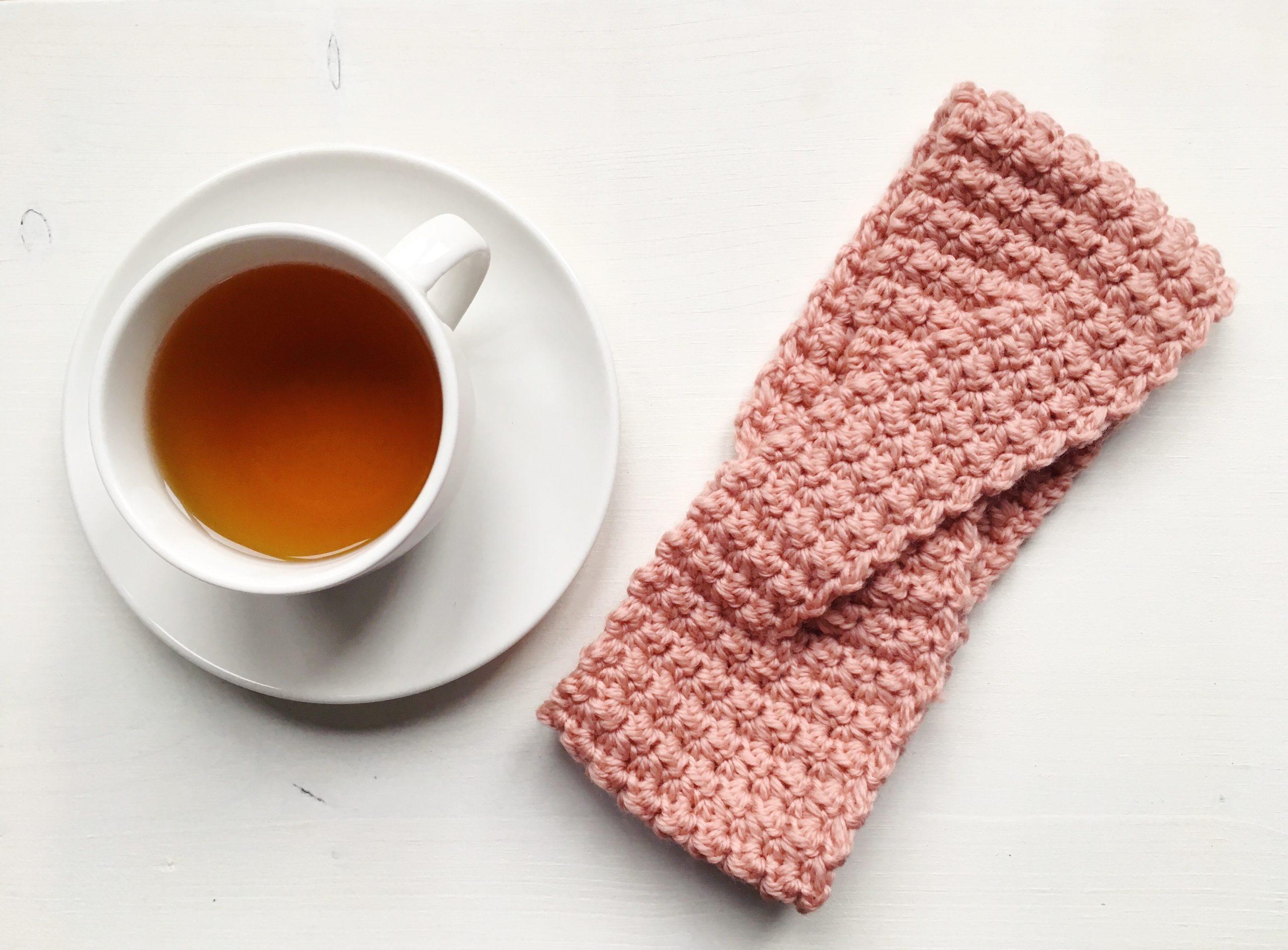 a muted pink crochet headband lay flat alongside a cup of tea on a white backdrop.