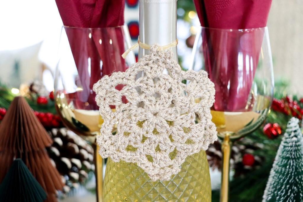 a close-up image of a crochet snowflake made in sparkly gold yarn attached to a bottle of wine on a decorated table for Christmas.