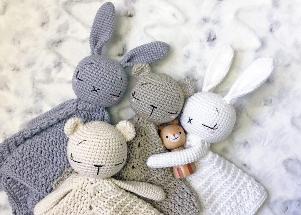 A foursome of crochet lovey blankets lay together, two are designed with bear heads and two with bunny heads. They are made in neutral shades of grey, tan and cream. 
