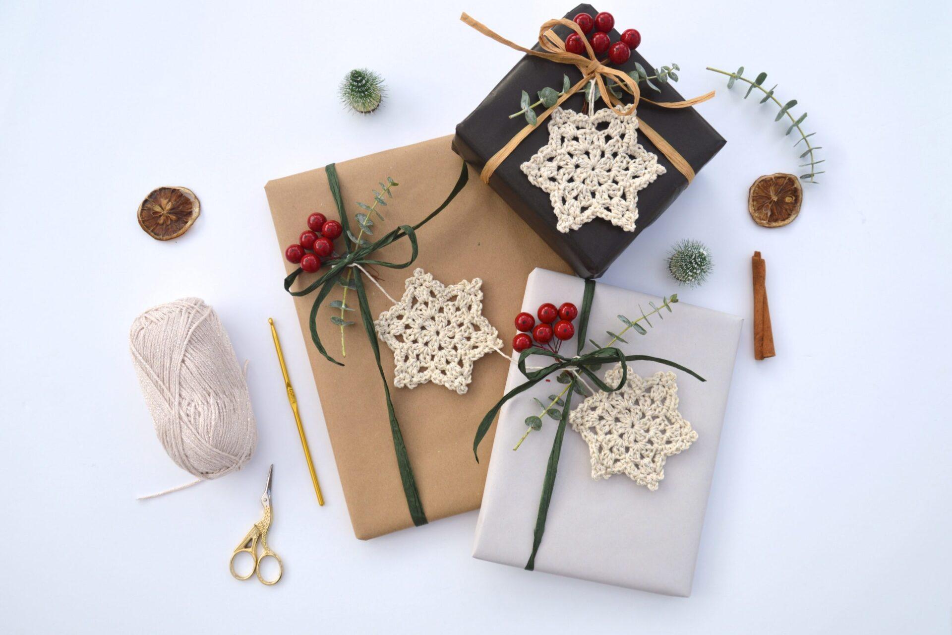 a flat lay image of three small gifts wrapped in neutral wrapping paper with a crochet snowflake in cream yarn attached to each of the presents along with foliage and berries.