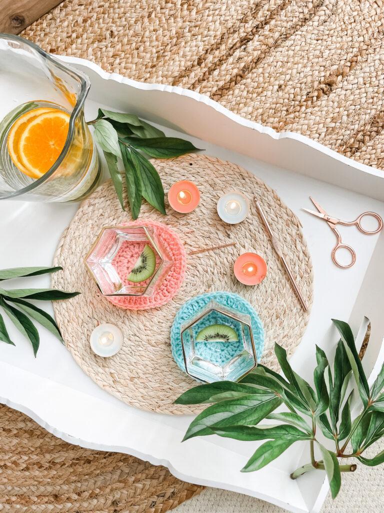a summery flat lay image of two crochet coasters in coral and blue shown in use with hexagon water glasses on them with slices of kiwi in the water. surrounding the glasses is a jug of water, tea light candles, foliage and crochet tools all on the same white tray sat in a jute rug. 