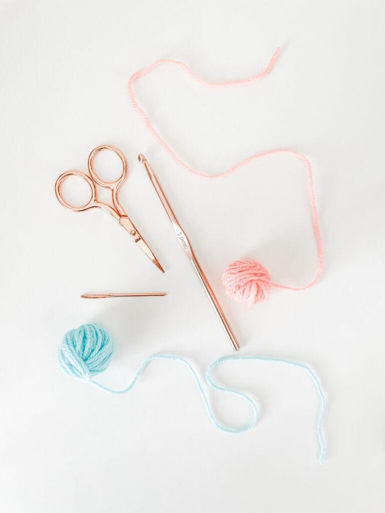 flat lay image of rose gold scissors, crochet hook, darning needle and two small balls of yarn, one blue, one orange on white background