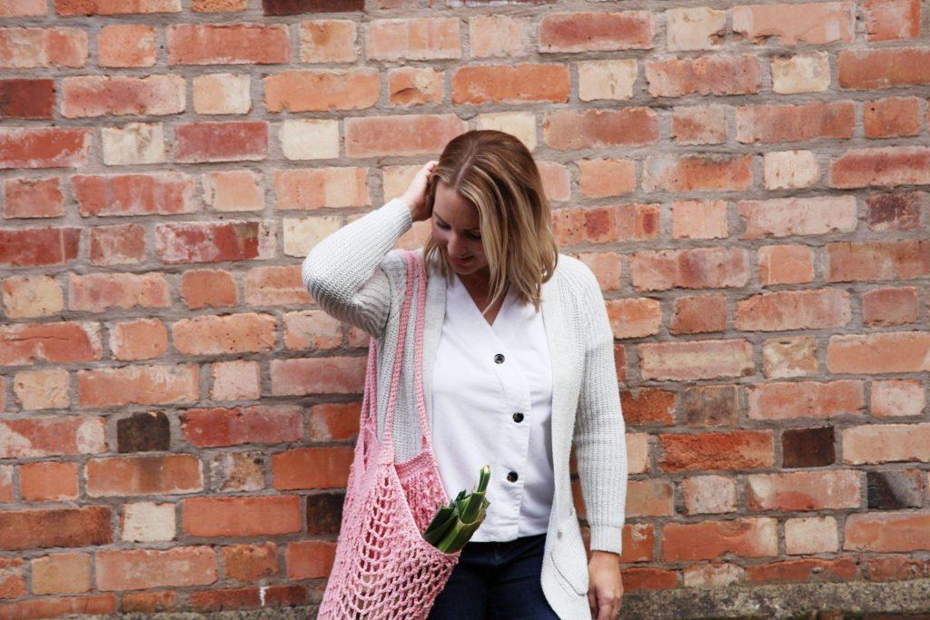 A blonde woman stands in front of a brick wall, she is smiling and has a pink crochet market bag on her shoulder. 