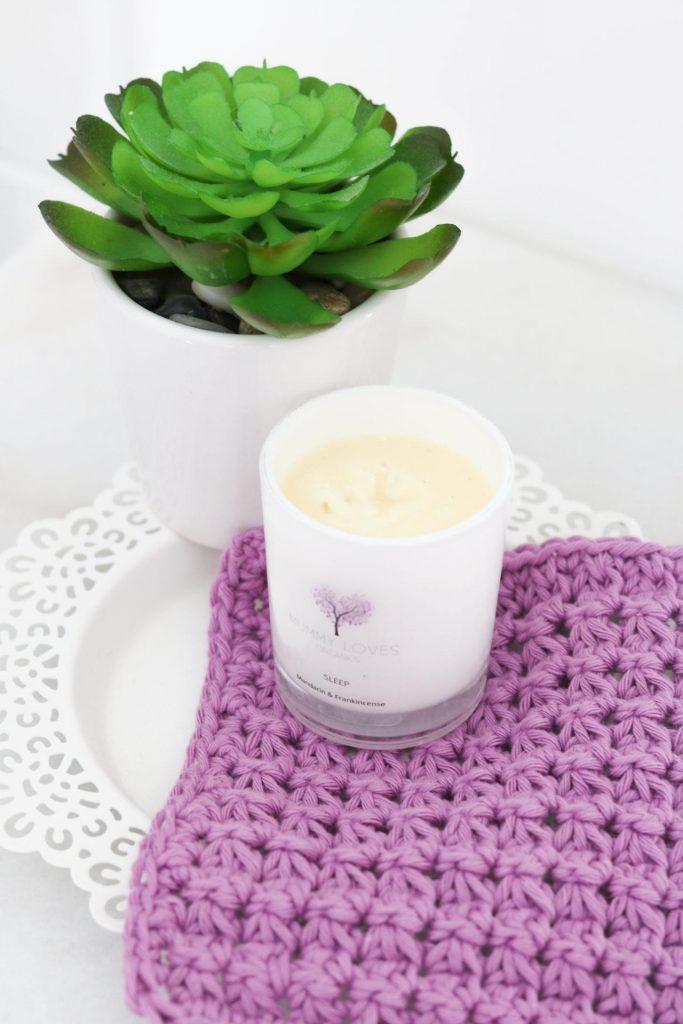 A purple washcloth made from crochet sits underneath a candle beside a cactus. 