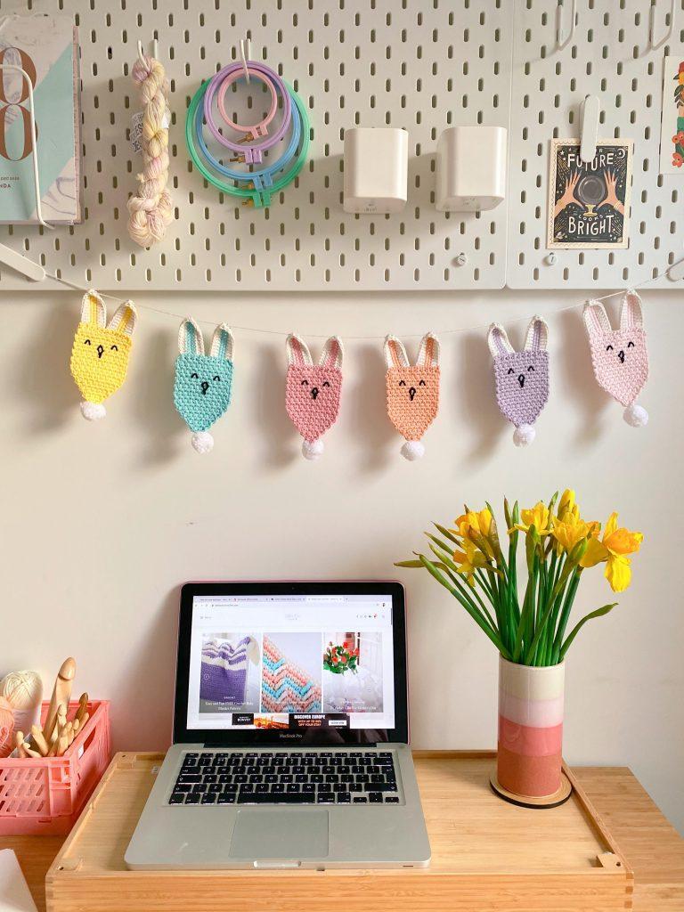 A crochet bunting string fashioned into bunny shapes is strung about a work desk with a laptop and bunch of daffodils sitting on it.