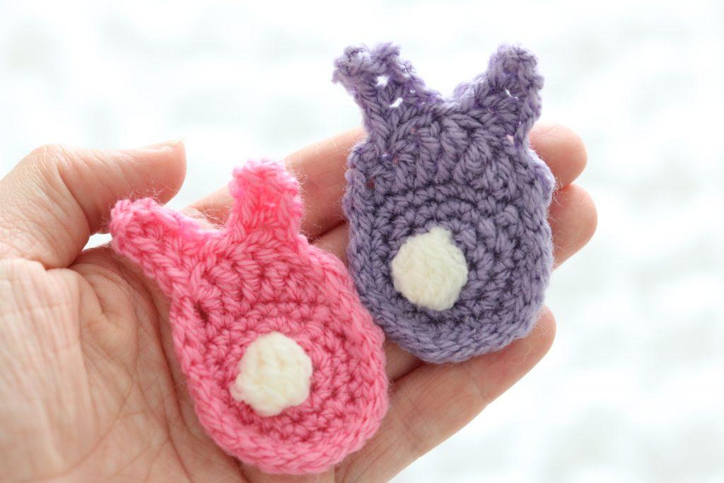 a pair of crocheted bunnies sits in a female hand, one is pink and one is purple. Both have white tails. 