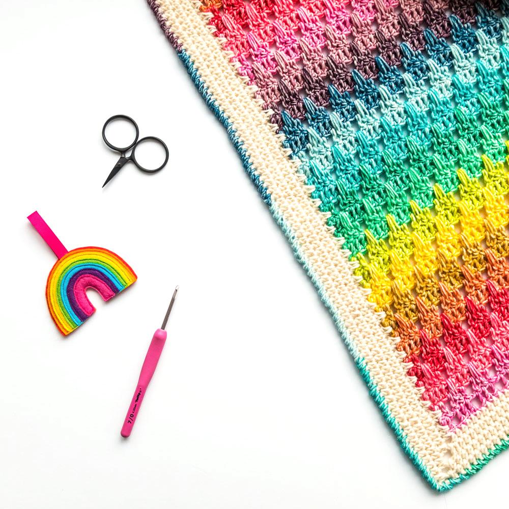 A rainbow-coloured blanket sits beside scissors, a pink crochet hook and a small felt rainbow