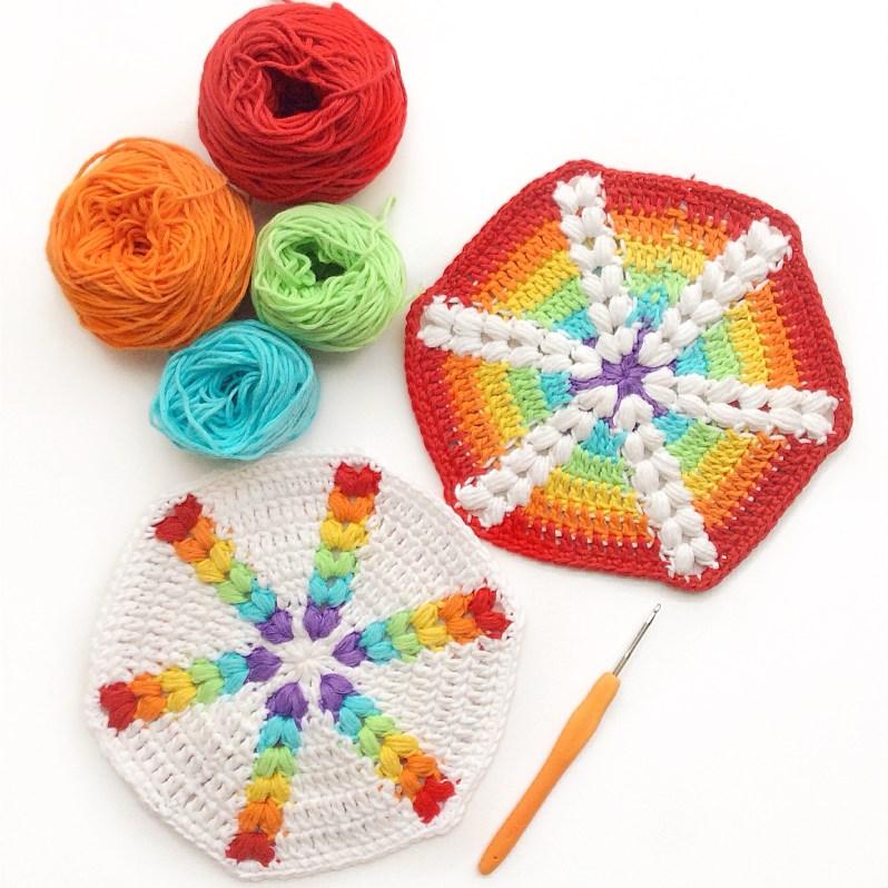 A pair of rainbow-coloured hexagons sit beside an orange crochet hook and four balls of yarn