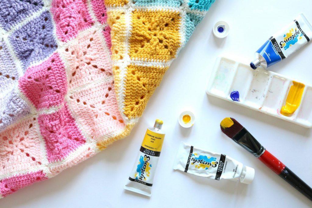 A pastel rainbow-coloured crochet blanket sits alongside colourful paints