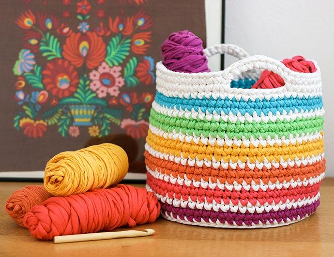 A white and rainbow-coloured crochet basket sits on a wooden worktop beside three skeins of yarn and a wooden hook