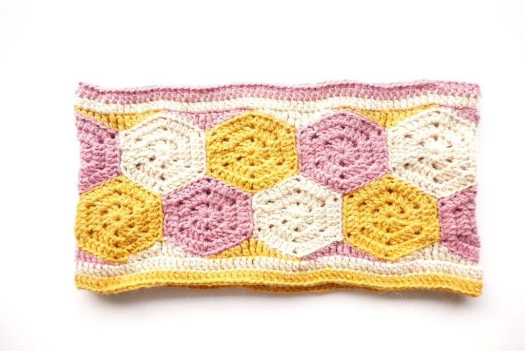 A cowl made up of crochet hexagons  in bright shades of white, pink and mustard sits on a white background. 