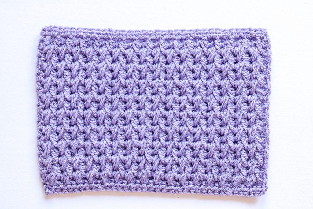 Feathered crochet stitch photographed from above on a white background. The swatch is in lilac yarn.