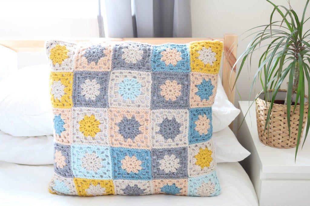 A crochet cushion made from granny squares in shades of grey, blue and yellow