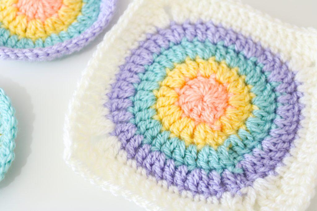 Flat lay image of a circular granny square. The circle is made up of four different colours: peach, yellow, teal and purple with a cream border squaring off the circular shape.