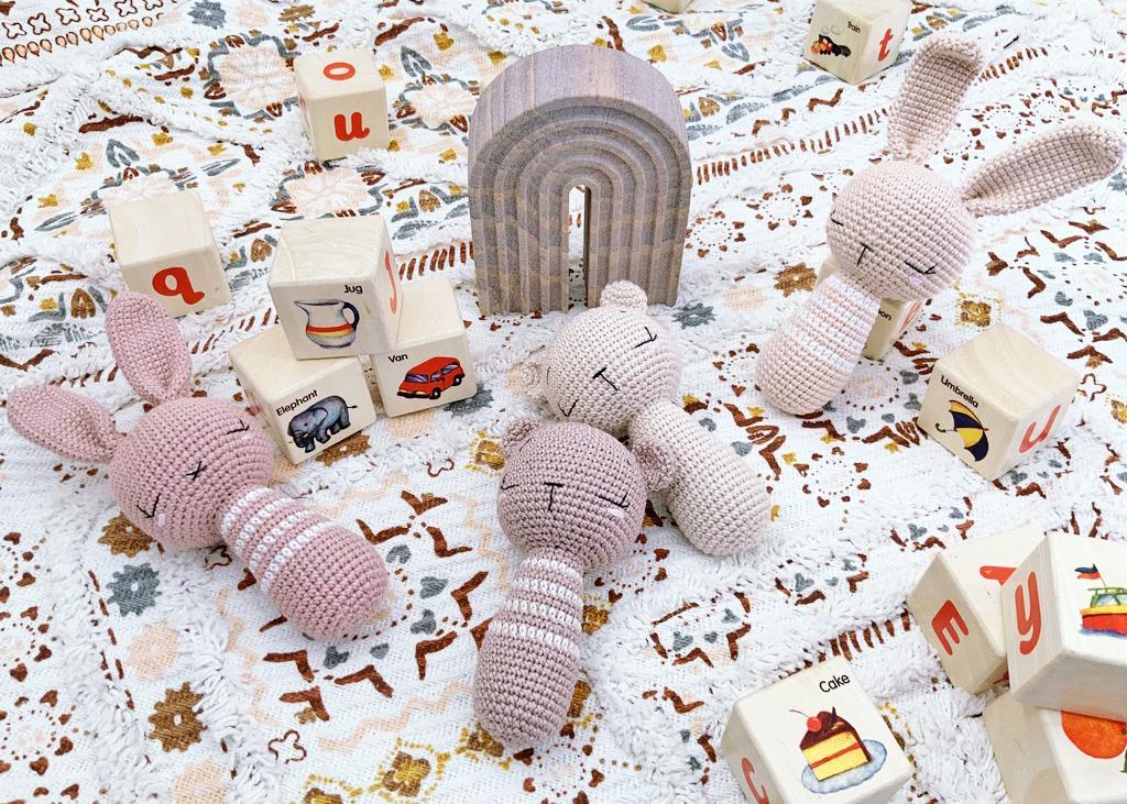 Four crochet rattle toys lay amongst wooden blocks on a playful fabric design. The rattles are varying shades of pinks and designed to look like sleepy baby rabbits and bears.