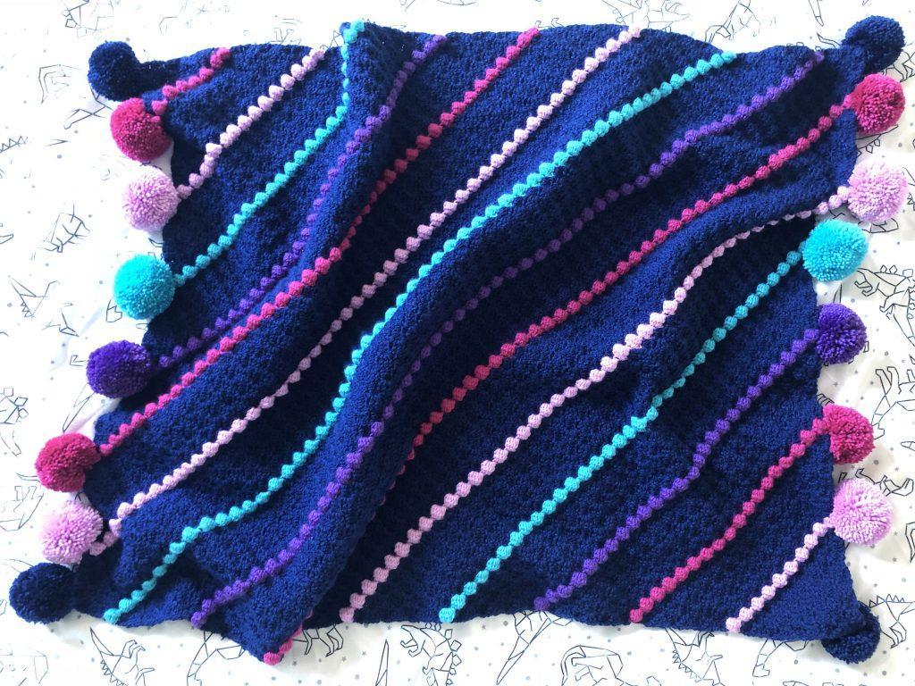 A navy blanket with rainbow stripes using the corner to corner method.