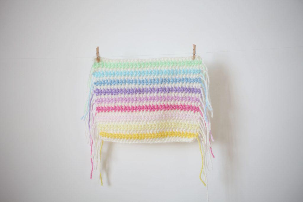 A swatch of half treble crochet in white and pastel rainbow stripes is show hanging against a white background.