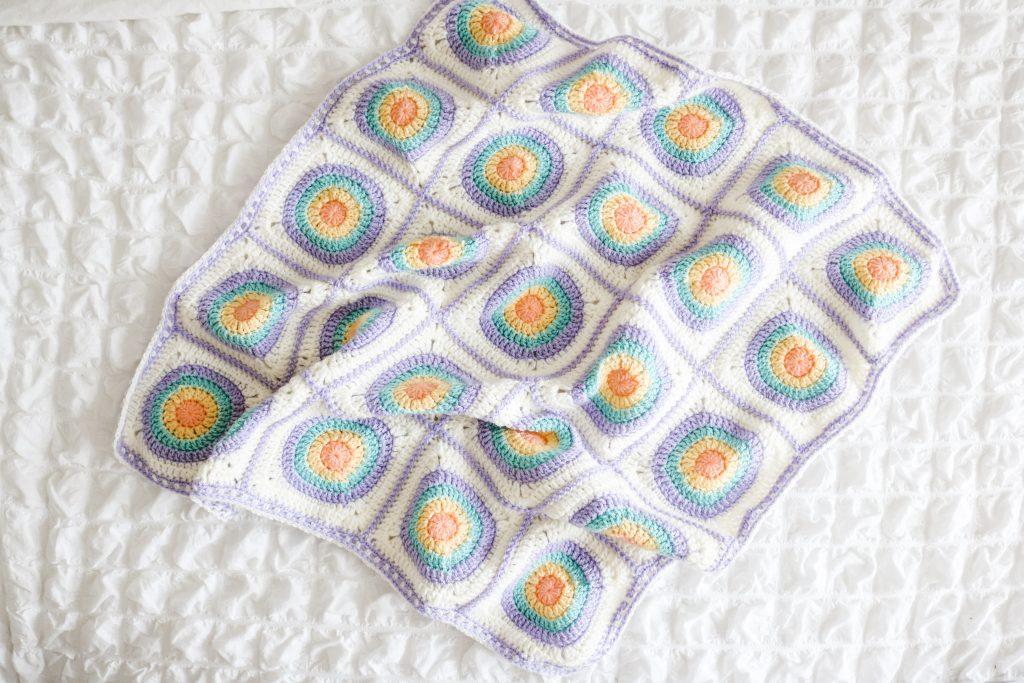 A completely circle of hope blanket in pastel shades of white, yellow, green, orange and purple sits on a white background.