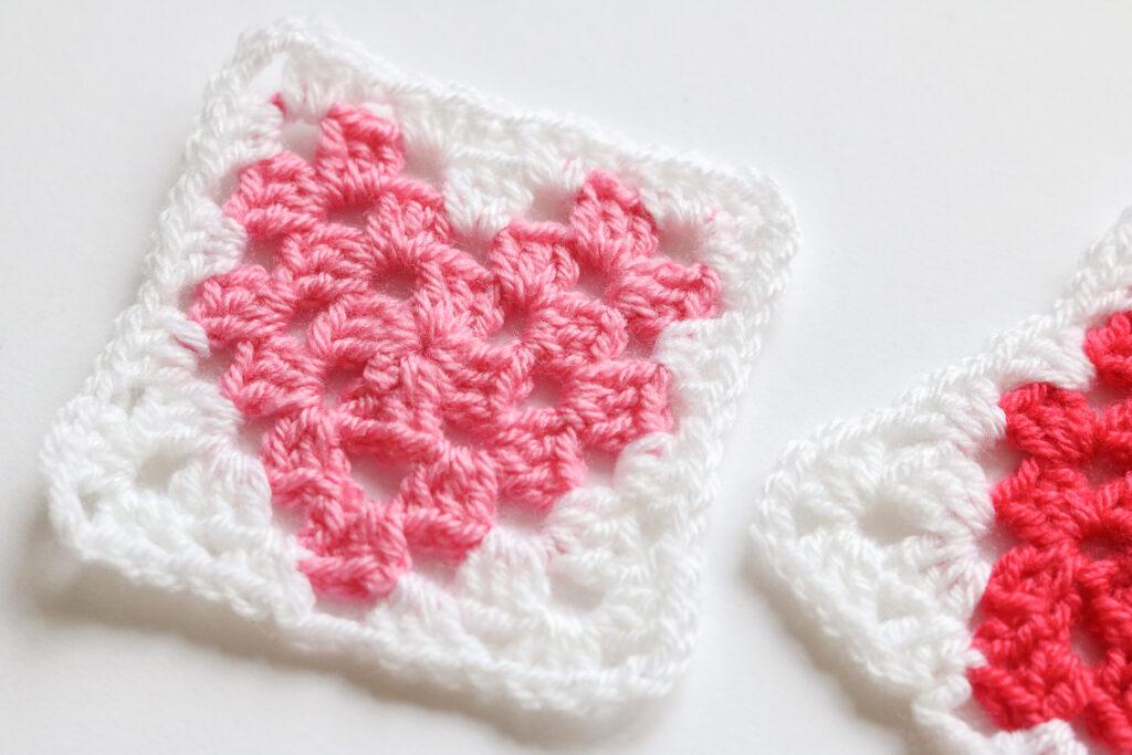 A pair of granny squares with a heart motif in the centre sit on a white surface.