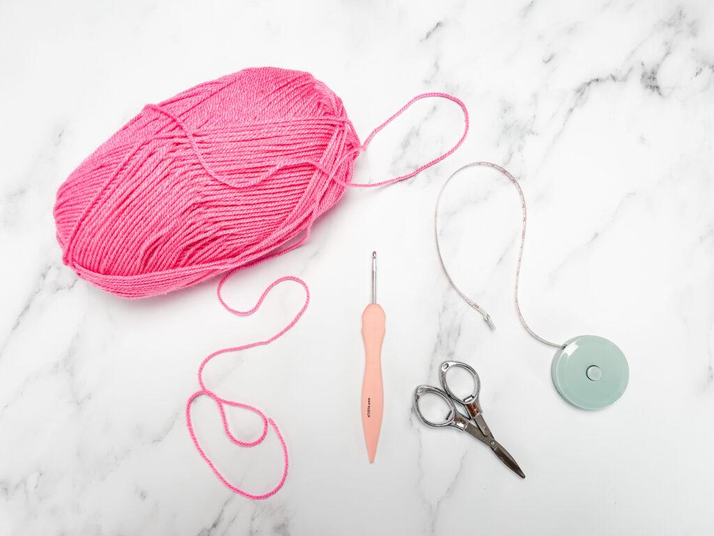 flat lay image on a marble backdrop including pink yarn, crochet hook, tape measure and scissors. 