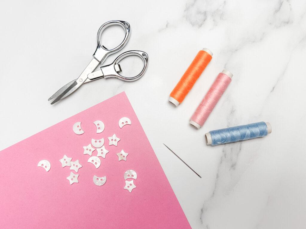 flay lay of three embroidery yarn spools in three colours; orange, pink and blue, sewing needle, scissors and white moon and star shaped beads on marble backdrop, the beads are lay on a pink piece of paper. 