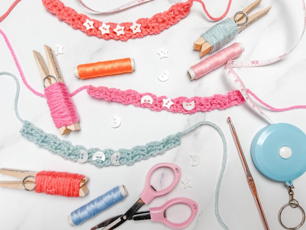 flay lay image on a marbled backdrop showing three crochet friendship bracelets in pink, orange and pale blue, yarn pegs, embroidery thread, scissors, crochet hook and tape measure.